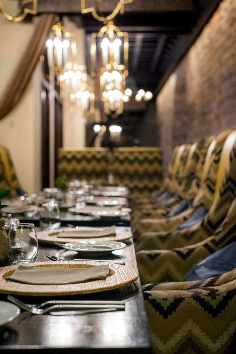 A long dining table set with white plates and glassware in a dimly lit restaurant with ornate chandeliers and patterned