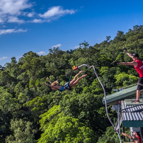 Person bungee jumping off a platform above a dense forest, with an instructor looking on and a clear blue sky in the background.