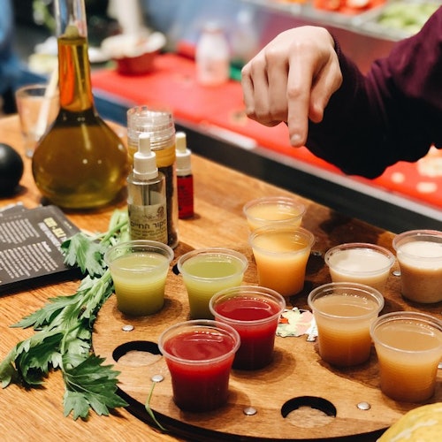 A person pointing at a wooden tray with various small cups of colorful juices, fresh herbs, and bottles on a counter.