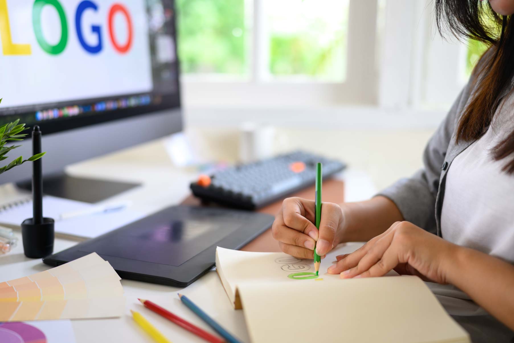 Grafik tworzący odręczne szkice koncepcyjne logo w notesie na stanowisku pracy z tabletem graficznym oraz monitorem wyświetlającym wektorowy projekt w procesie kreacji brandingu