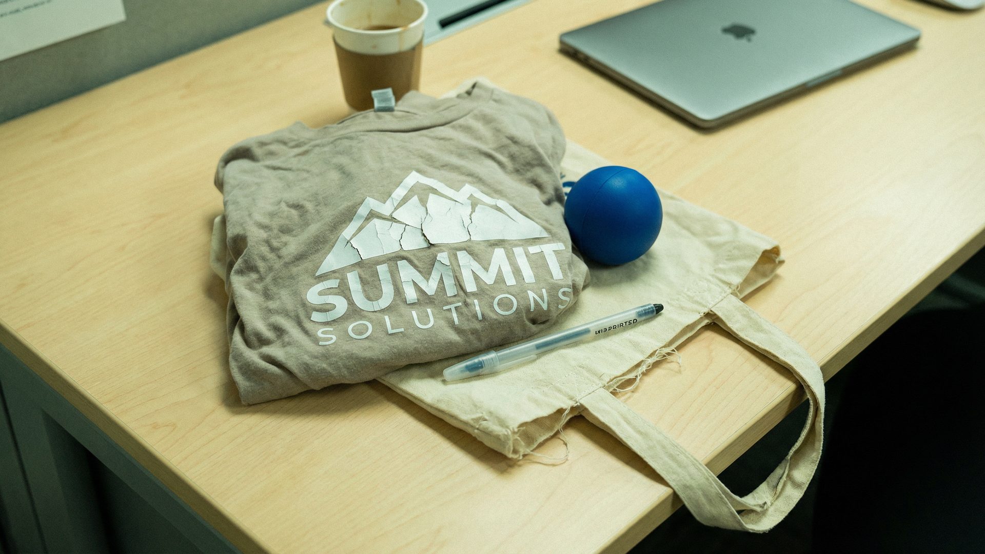 A desk covered in cheap, low-quality promotional merchandise — a t-shirt with a cracking logo, a flimsy tote bag, a stress ball, and a generic pen — representing the disappointment of traditional swag vendors.
