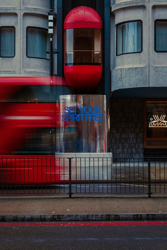 Fotografia uliczna przedstawiająca budynek w Londynie z czerwonym balkonem, rozmazany jadący czerwojy autobus oraz witrynę z napisem "SET YOUR SPIRIT FREE"