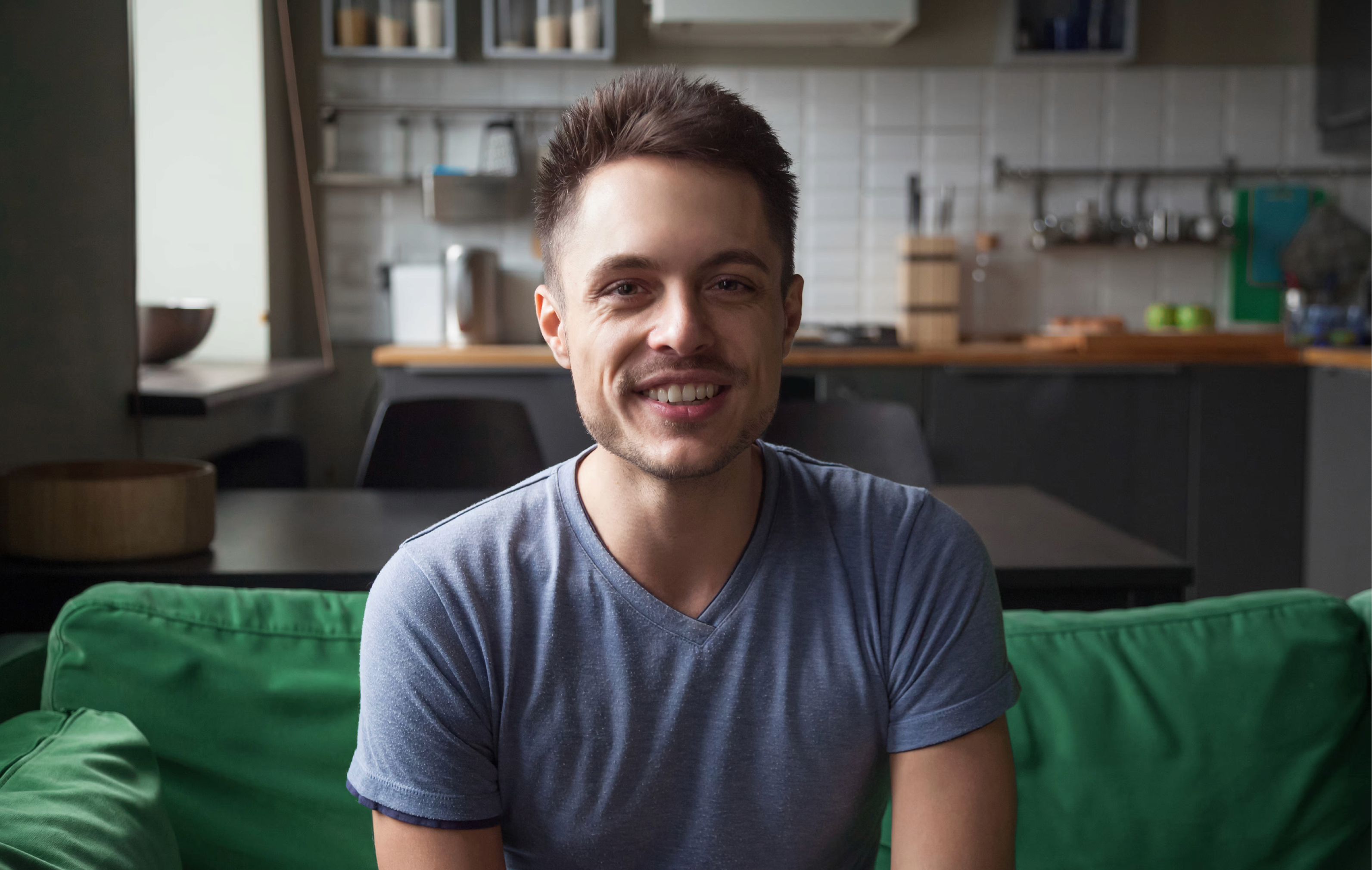 Smiling man on a video call, seated on a green sofa in a home kitchen, looking at the camera.