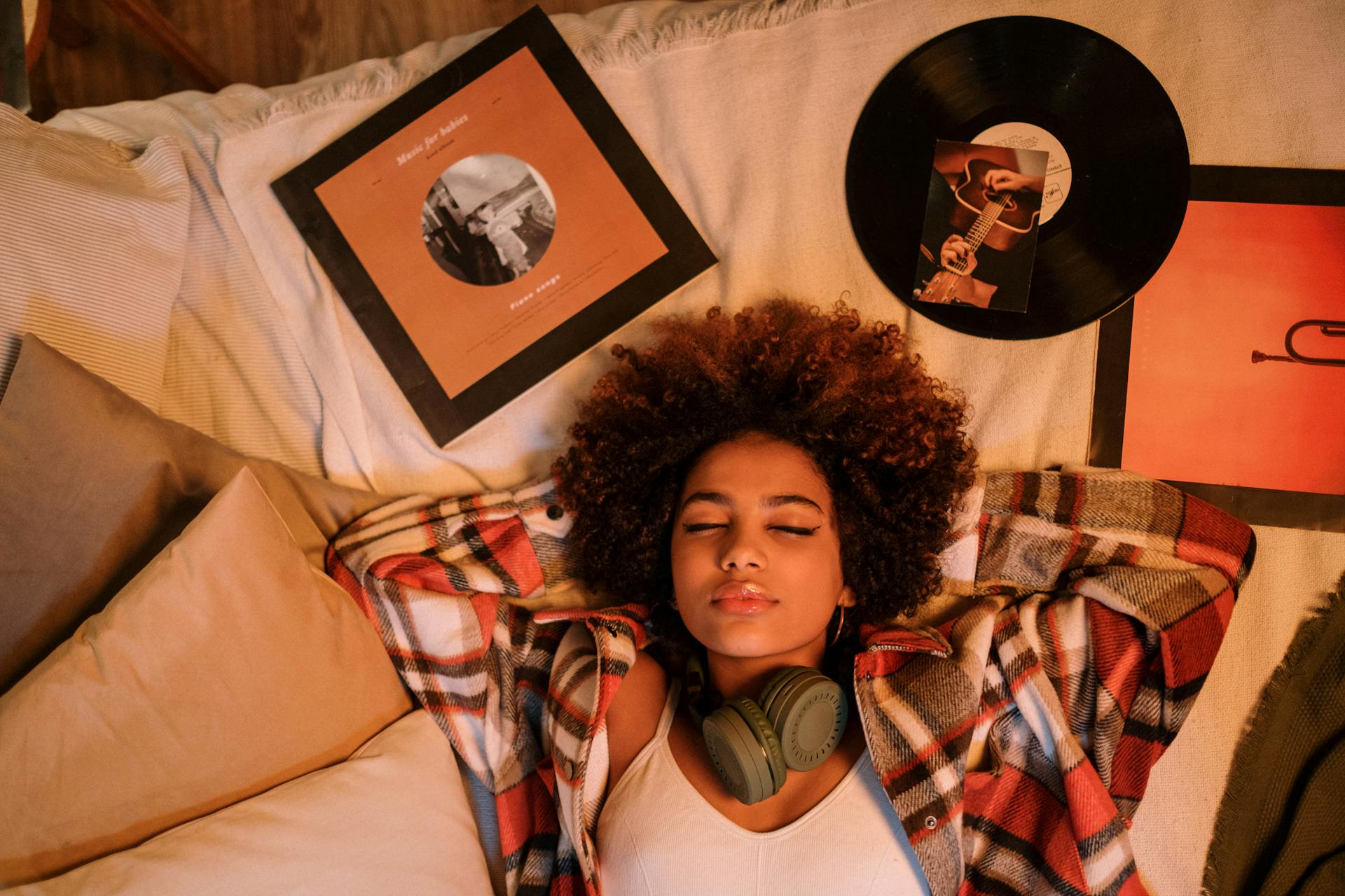 A top-down view of a young woman with curly hair lying on a bed with her eyes closed, surrounded by vinyl records and album covers.