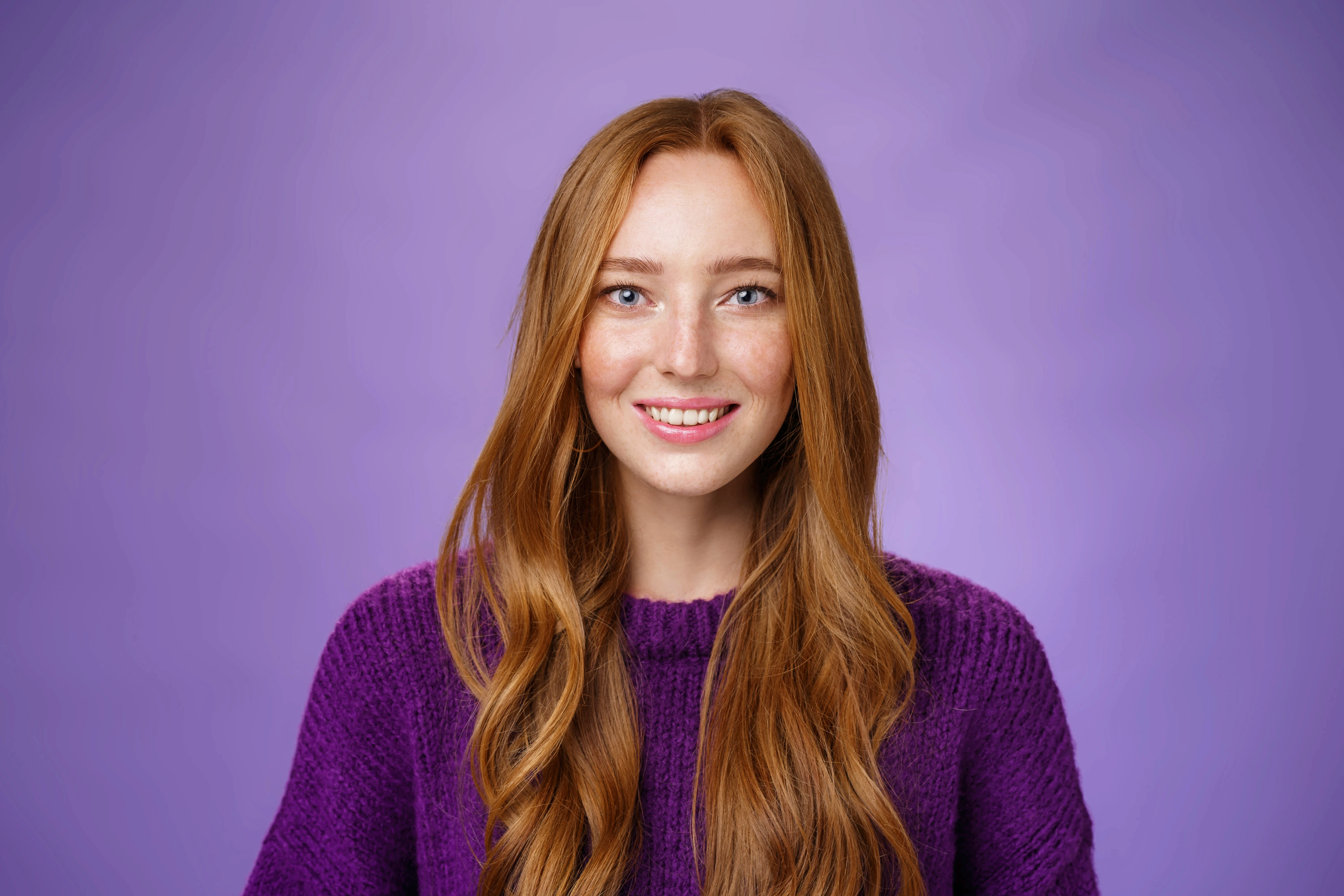 A person with long red hair smiles warmly against a soft purple background, wearing a purple sweater. The atmosphere is cheerful and inviting.