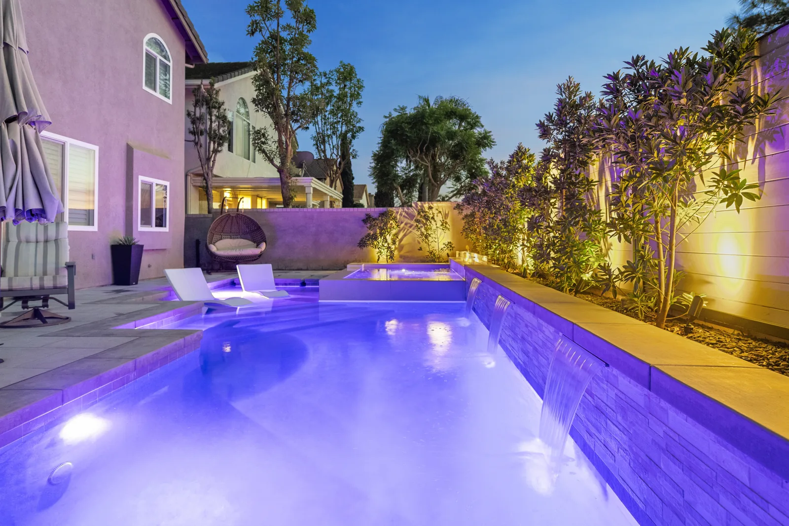 Pool, spa and waterfalls An illuminated pool beside a home, with a raised spa, in-water loungers, waterfalls and a paved deck.
