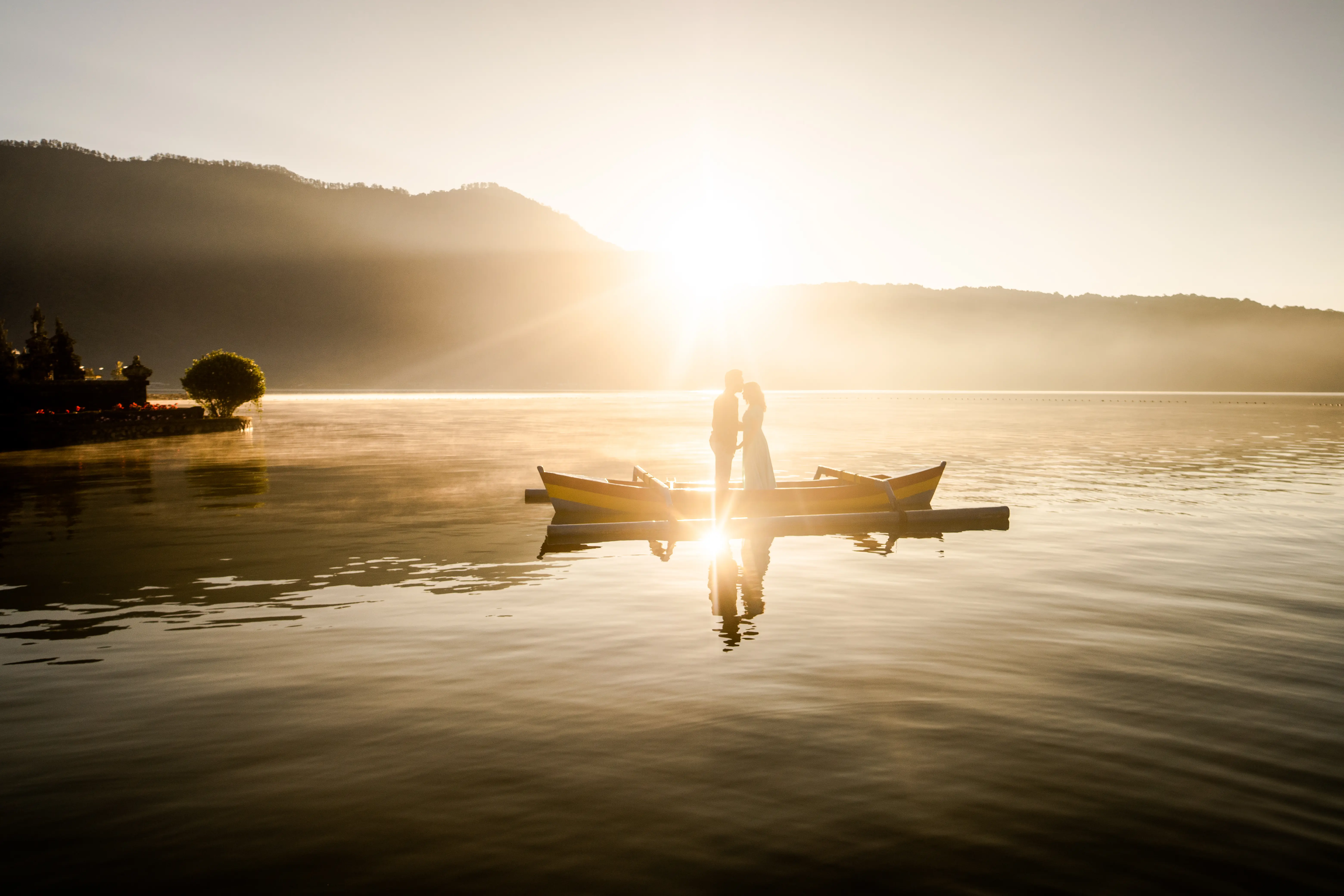 A couple in a boat glide across a still lake at sunrise in Bali, with soft light illuminating the peaceful morning scene. Fine Art Wedding Destination Photography in Hyderabad by Out of The Blues, photo taken in Bali.