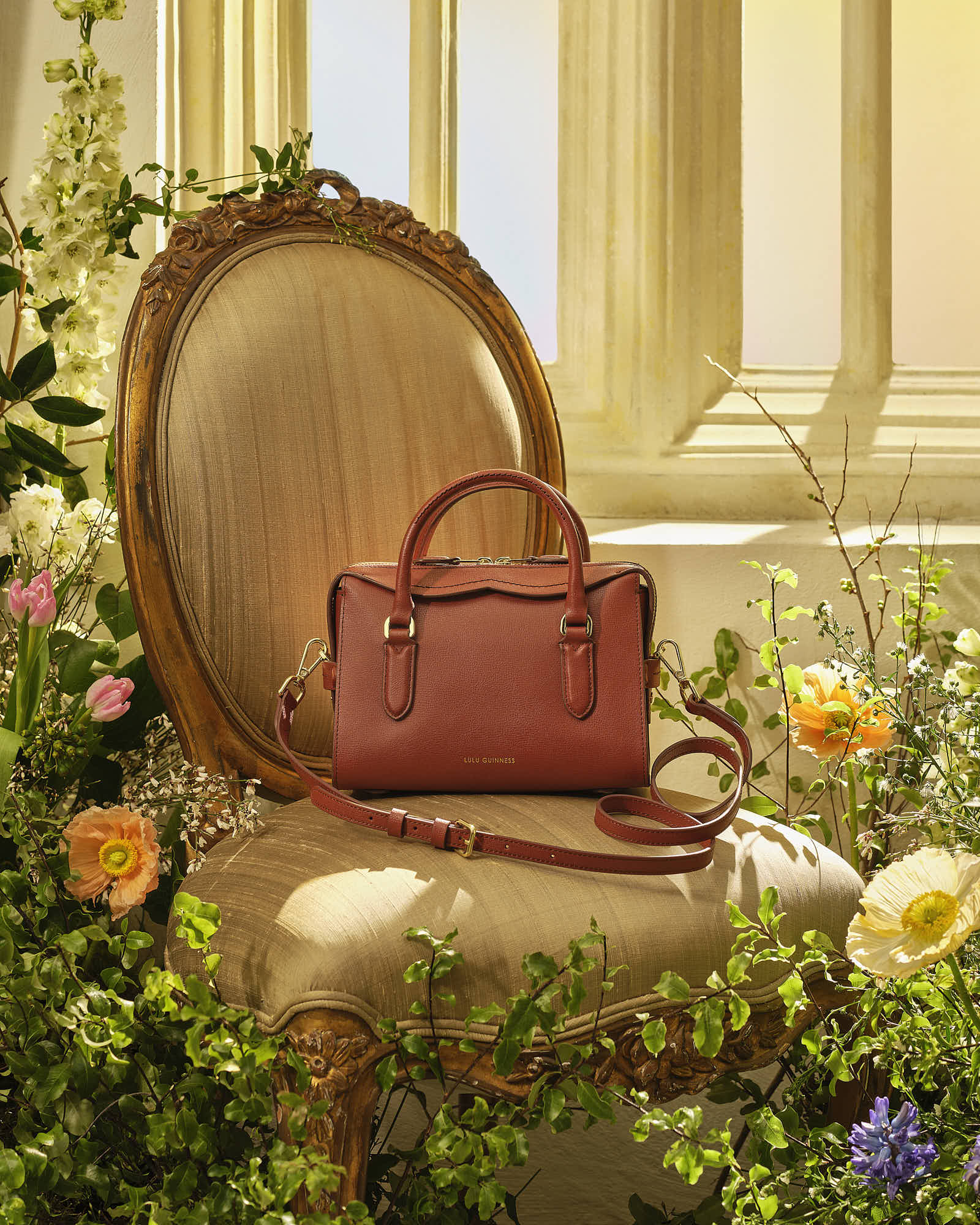 A stylish red handbag placed on a luxurious chair surrounded by colorful flowers and soft, warm lighting.