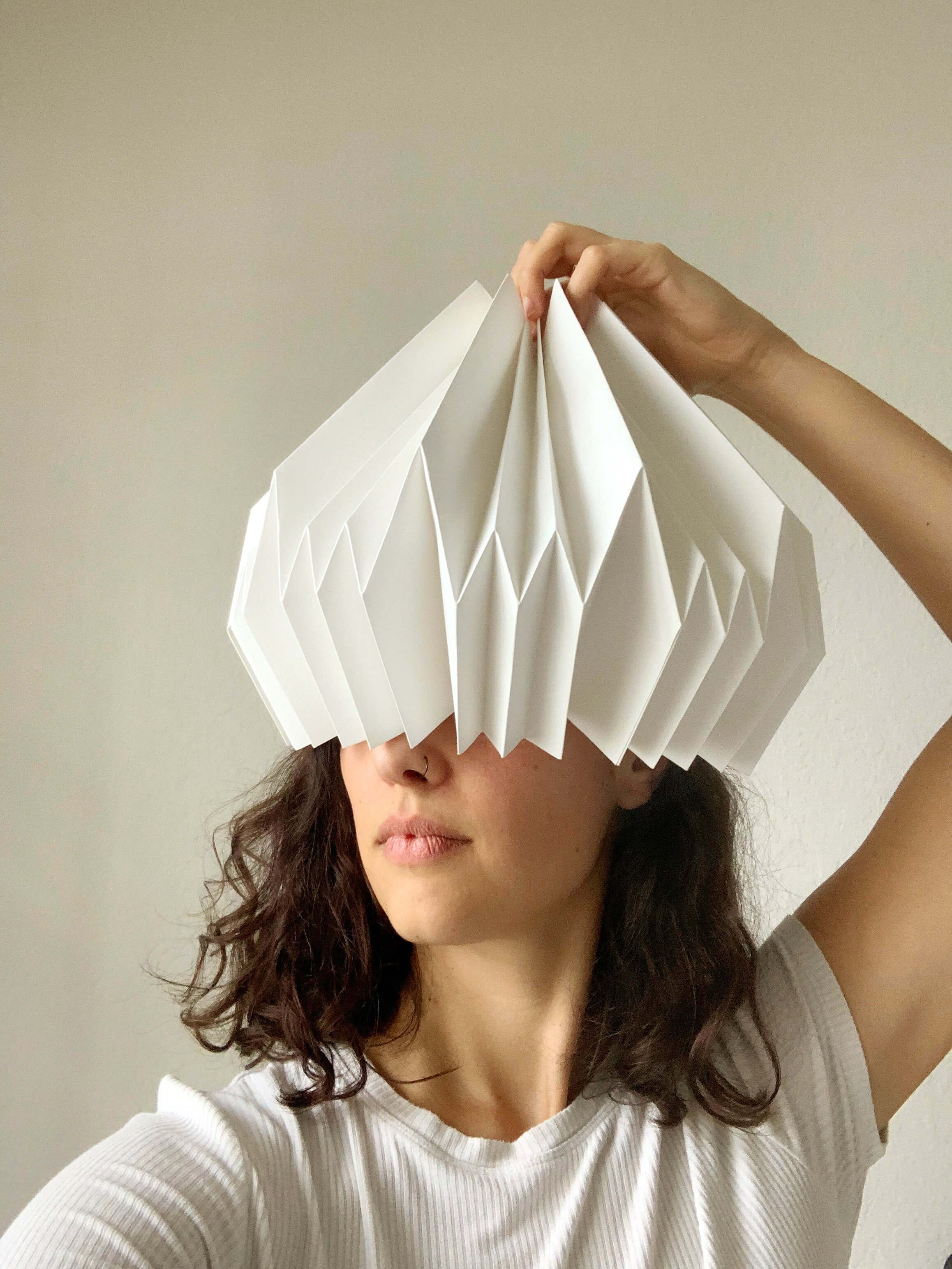  Person holding a folded white paper sculpture over their eyes like a visor, partially covering their face, creating a playful and artistic composition against a neutral background.
