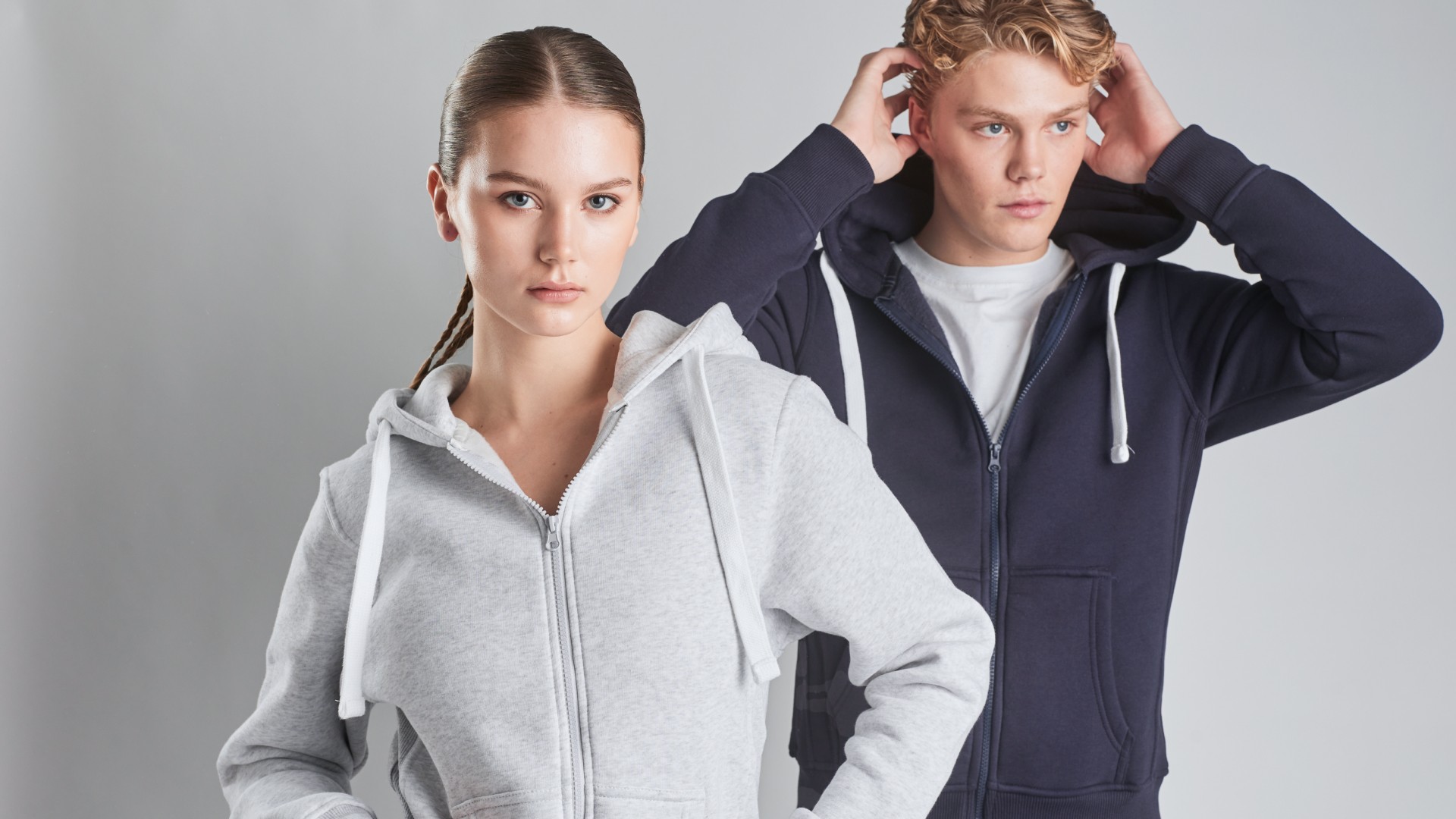 Two young people, one male and one female, wearing light grey and dark navy ziphoodie in a studio setting.