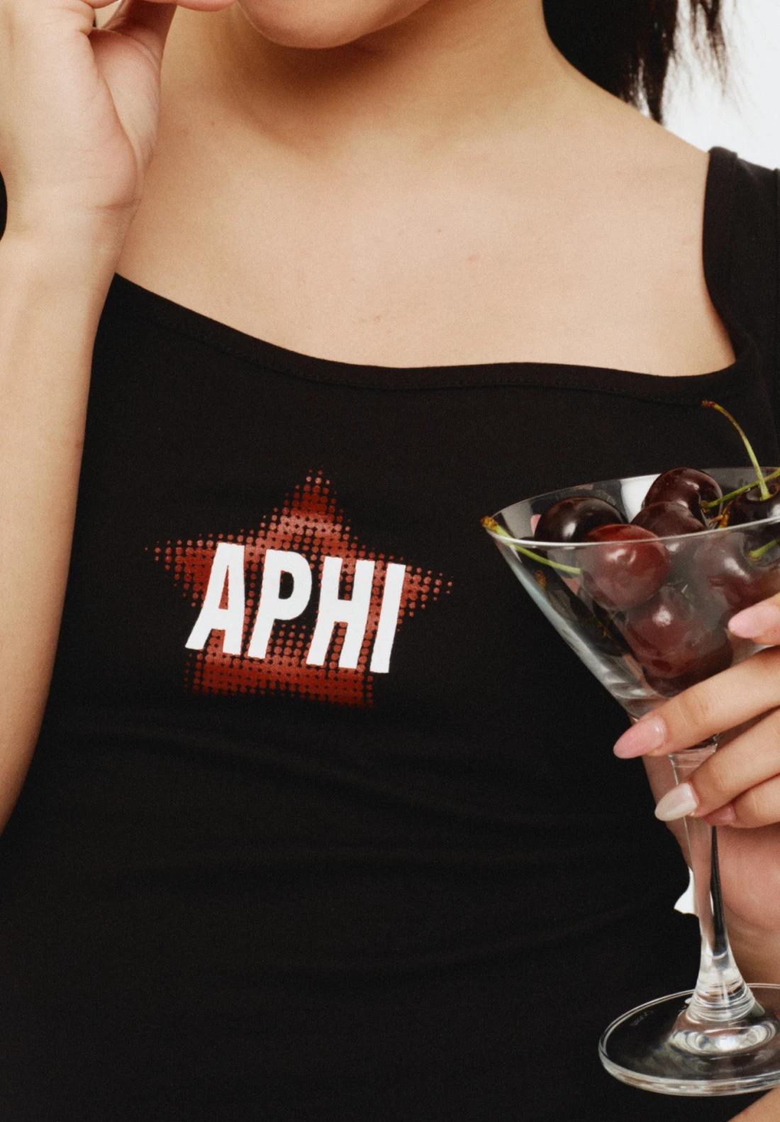 Close-up of a black tank top with “APHI” graphic, holding a martini glass filled with cherries
