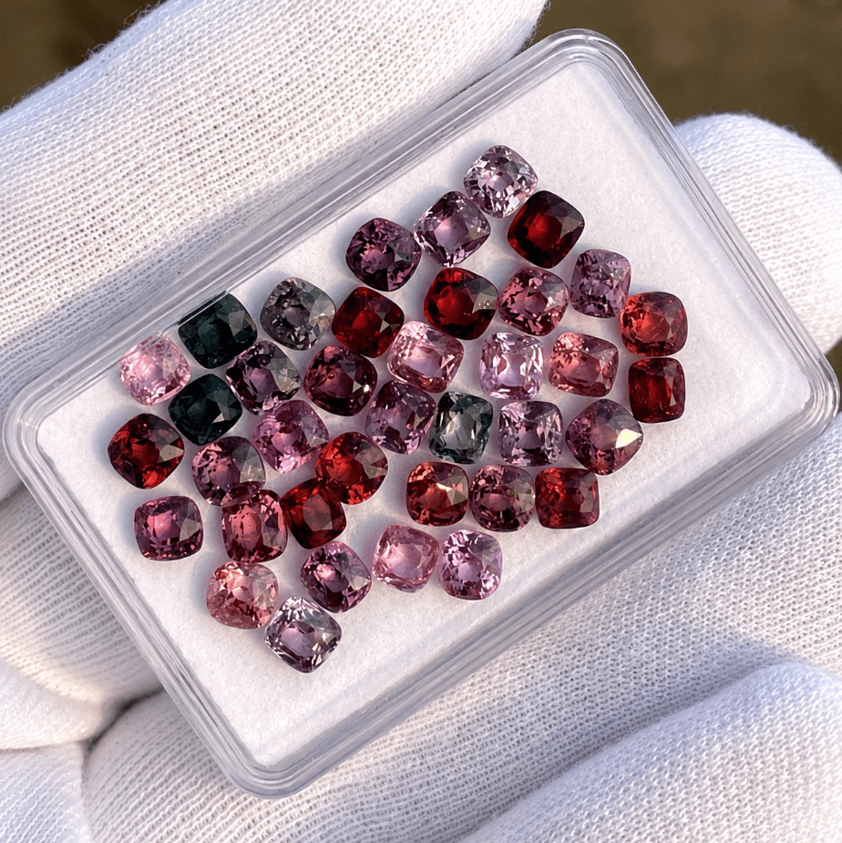 Assortment of dark coloured faceted Lab Grown Spinels in a box held by white gloves.