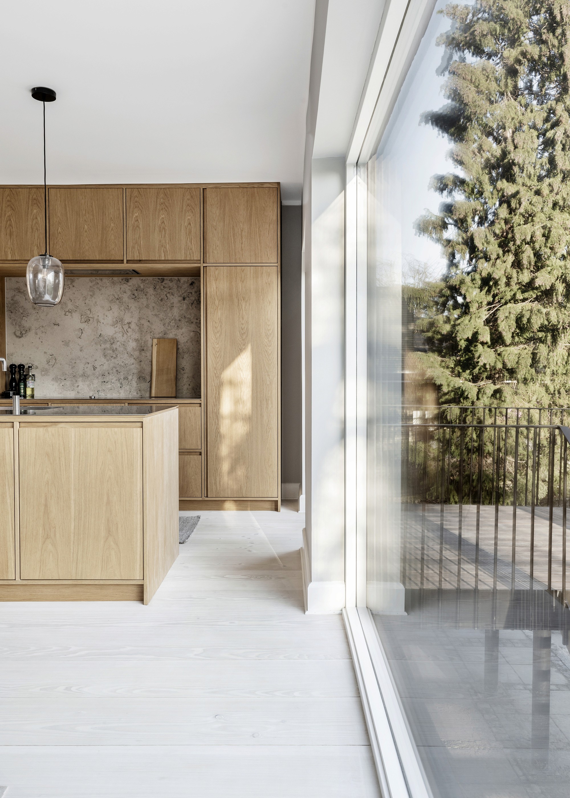 Moderne køkken med lyse træskabe og store vinduer med udsigt over træer. Rummet er lyst og minimalistisk og tilbyder en rolig atmosfære.