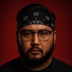 A woman with blonde bob hair in a grey coat and beige turtleneck stands against a neutral backdrop. She has a serious expression, conveying elegance.