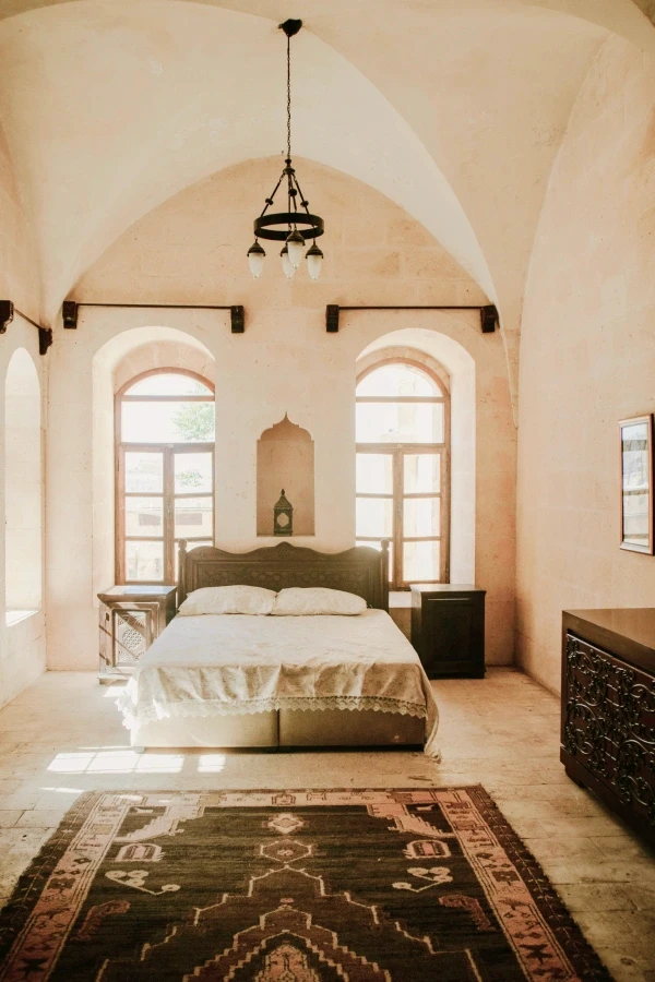 Bright afternoon sunlight fills a rustic stone room through tall panelled windows, illuminating the floor next to a large bed and dark wood chest of drawers.