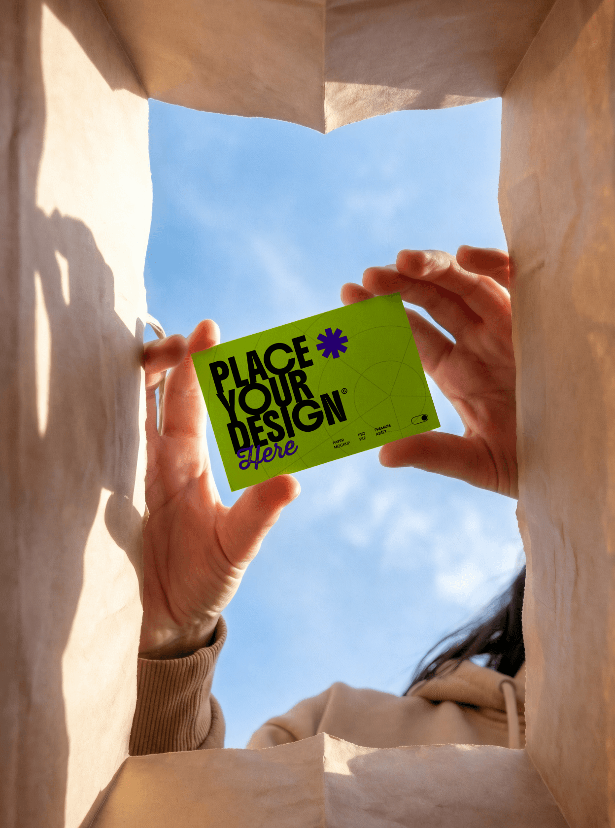 A unique low-angle shot from inside a brown paper bag looking up toward a blue sky. Hands are shown holding a lime green business card mockup with "Place Your Design Here" text, framed by the edges of the bag.