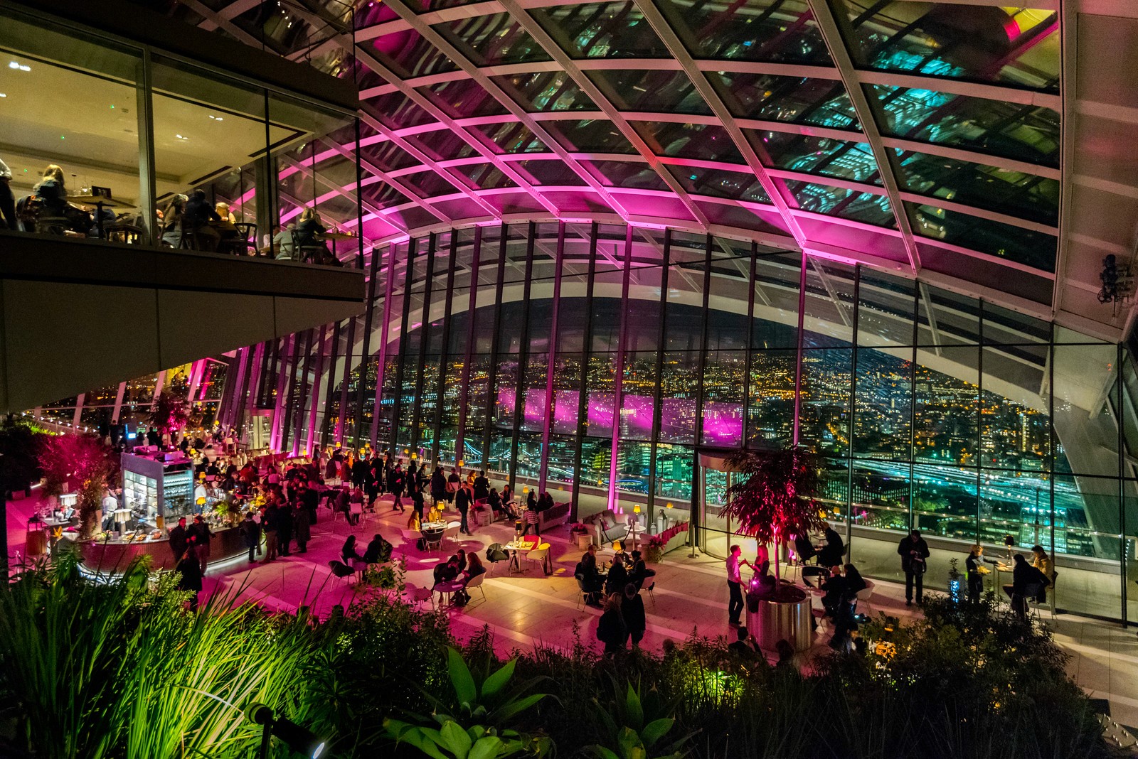Crowded indoor lounge with pink and blue neon lighting under a glass vaulted ceiling with people sitting at tables.