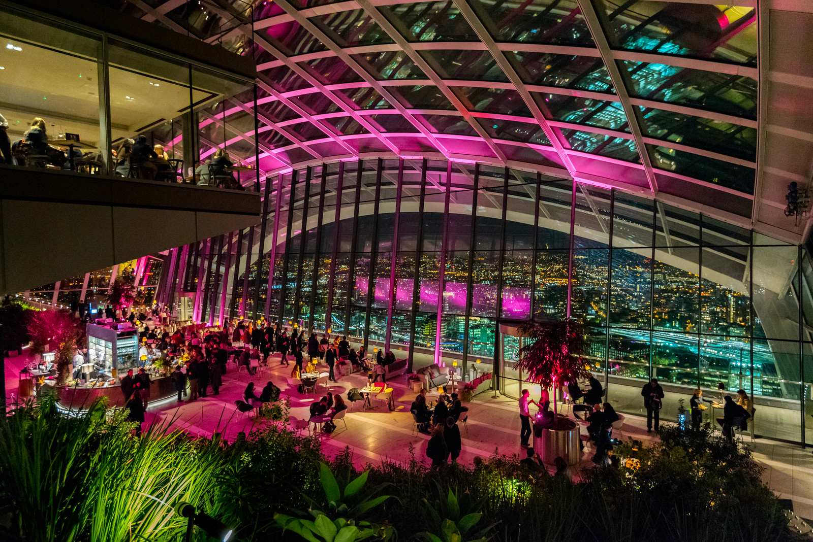 Crowded indoor lounge with pink and blue neon lighting under a glass vaulted ceiling with people sitting at tables.