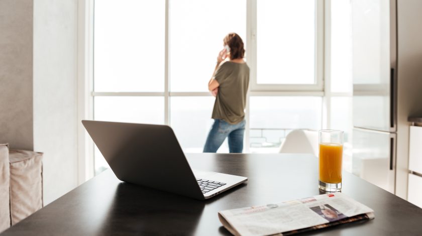 A modern home office setup features a sleek laptop on a black table next to a glass of orange juice and a folded newspaper, while a person stands by large, sunlit windows in the background.
