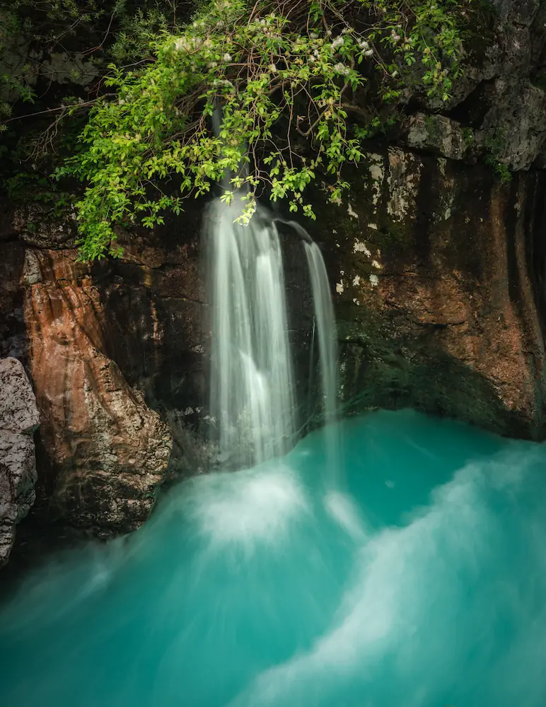 A long-exposure photo of a small waterfall flowing into the vibrant turquoise water of the Soča River gorge in Slovenia, surrounded by dark mossy rocks and overhanging green foliage.