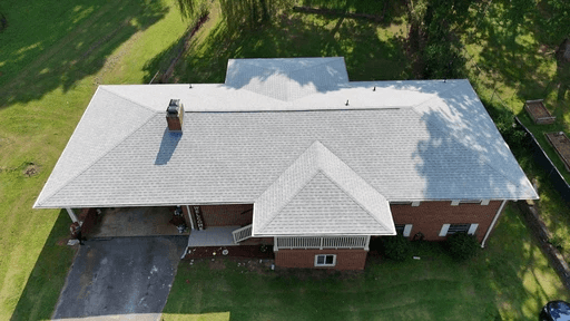 Aerial view of a single-story house with a gray roof, surrounded by green grass and nearby structures.