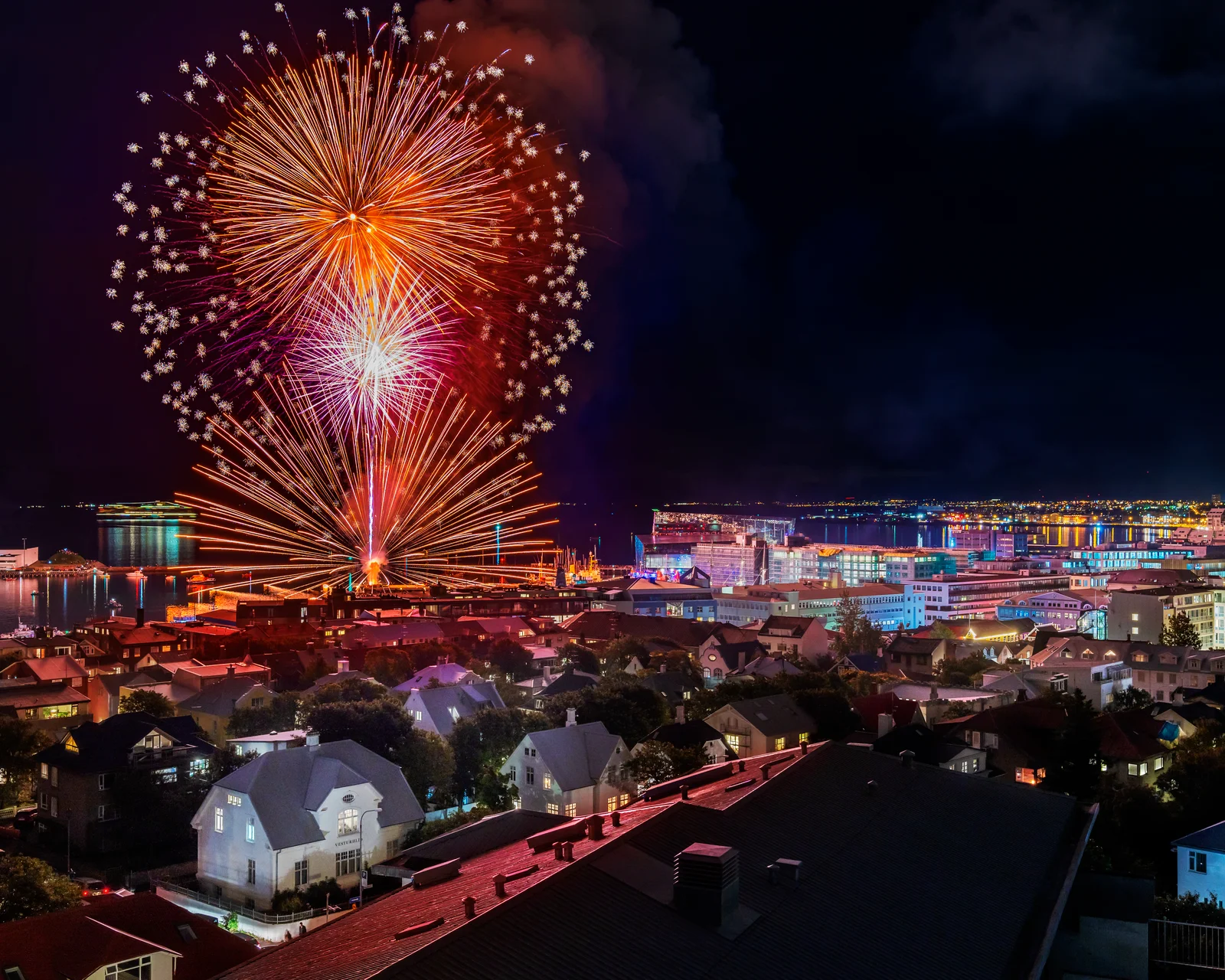 Vibrant orange, red, and white fireworks explode in the night sky directly above the harbor during Reykjavík Culture Night. The illuminated cityscape, residential rooftops, and harbor front are visible below against a dark sky and ocean backdrop.