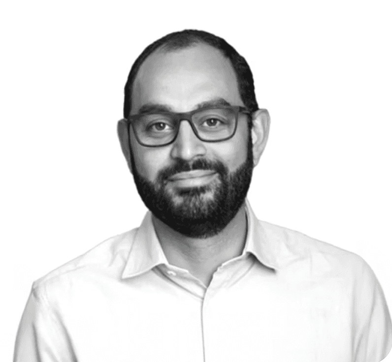 A focused woman with long red hair is seated at a desk, wearing a black blazer and dark blouse. She intently looks at camera, smiling slightly, with an open laptop in front of her. The setting is a warm and serene office setup in the background. Soft daylight filters through, creating a calm atmosphere. The composition captures her in a frontal profile with a clear emphasis on her concentration and the serene indoor setting.