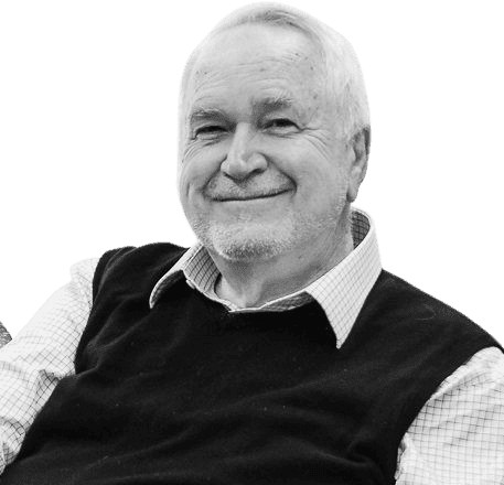 A black-and-white photo of an elderly man with a friendly smile, wearing a collared shirt under a dark sweater vest. He has a light beard and short, neatly combed white hair, giving him a warm and approachable appearance. The background includes office equipment and windows, suggesting a professional or academic setting.