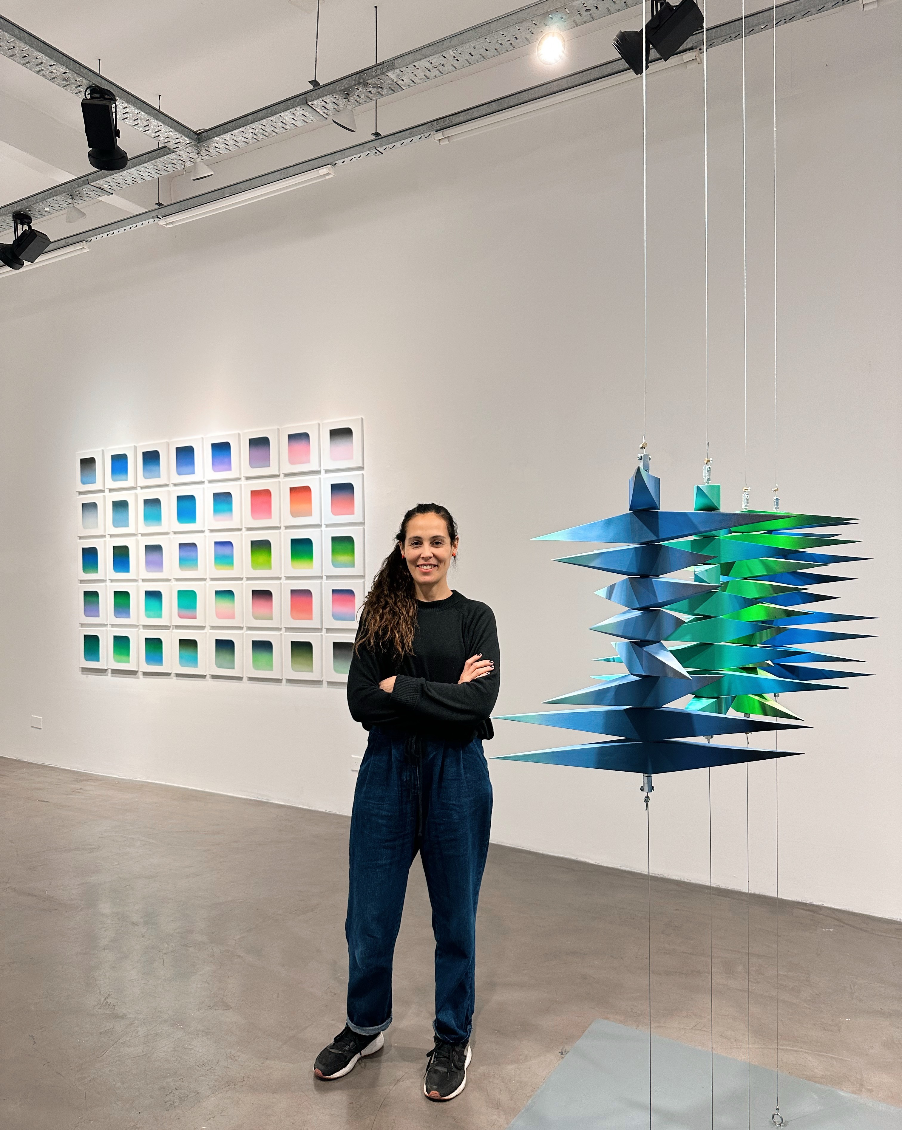 Captura a la artista sonriente frente a una exposición artística en la galería Gachi Prieto. Detrás de ella, se destacan dos instalaciones: una serie de cuadros con gradaciones de colores y una escultura colgante geométrica en tonos azul y verde, representando la intersección del arte geométrico y la interacción de colores.