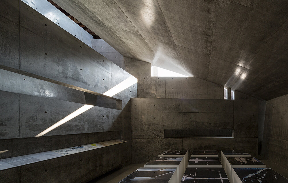 Concrete interior of Ando Museum with dramatic natural light entering through narrow openings in the angular exposed‑concrete ceiling, illuminating low display tables and emphasizing Tadao Ando’s minimalist architectural design.