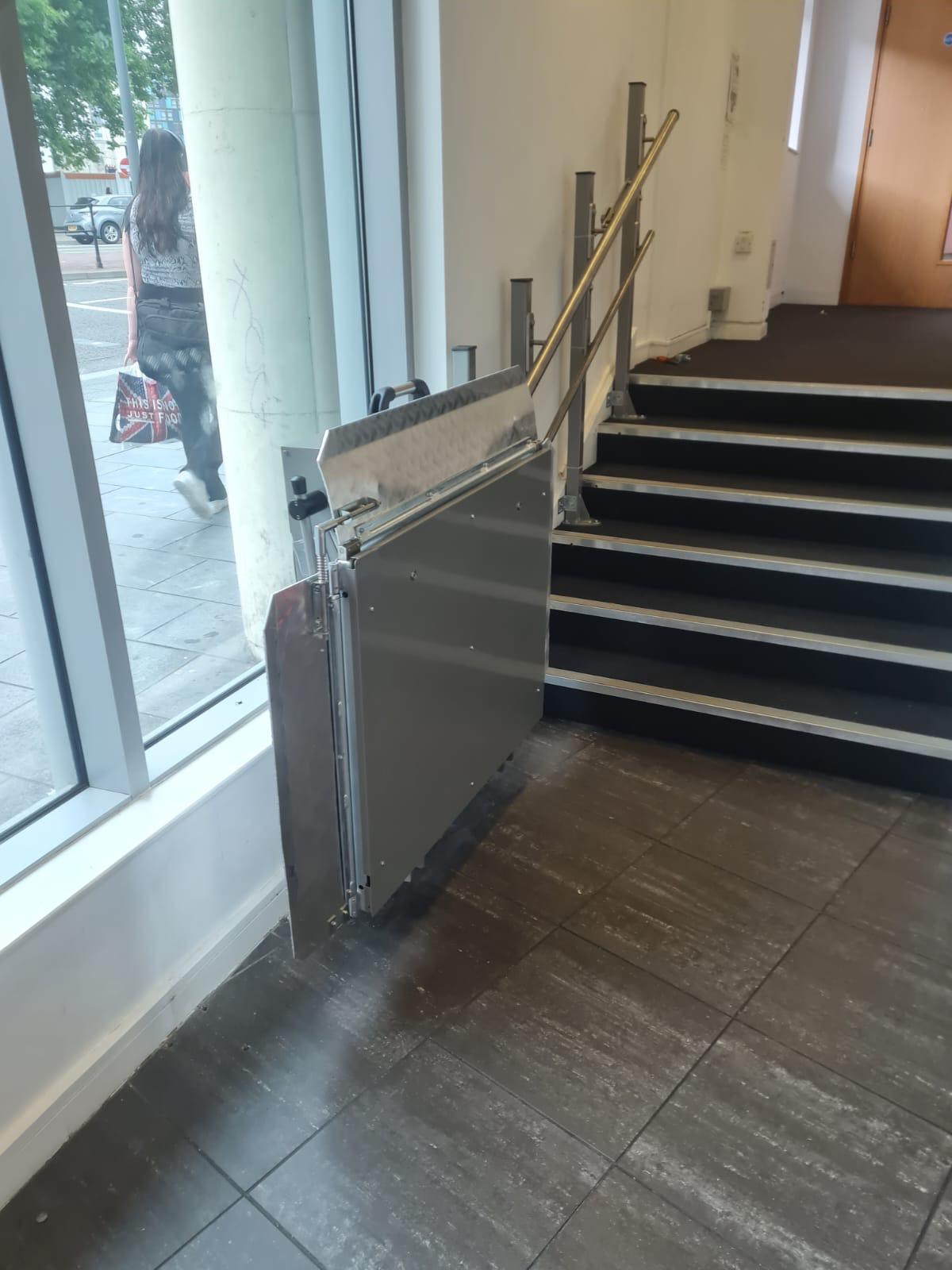 Straight incline platform stairlift in fully folded parked position at lower landing — stainless steel platform folded flat beside commercial staircase leaving stairs completely clear