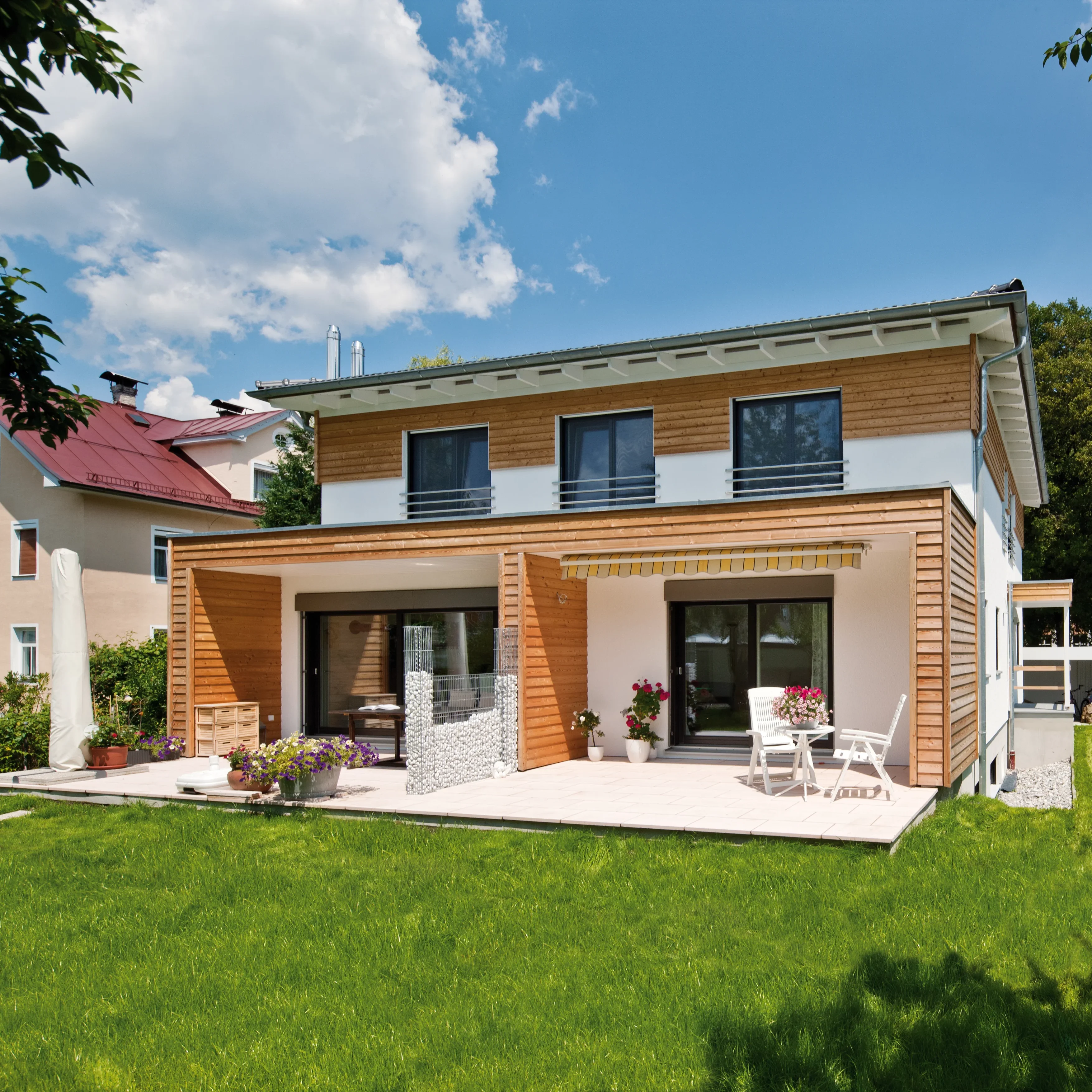 Modernes Haus mit viel Holzverkleidung, großzügiger Freifläche mit Terrassenmöbeln und blühenden Blumen im Garten.