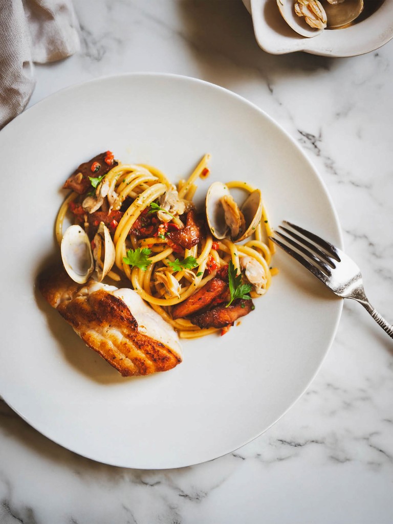 Overhead view of a plate of spaghetti with clams, a piece of seared fish, and a fork.