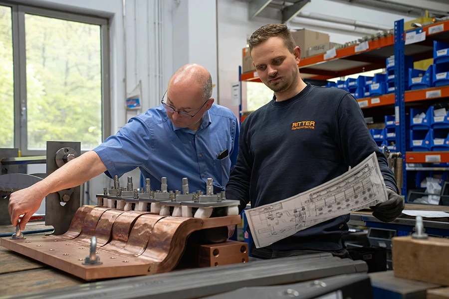 Zwei Mitarbeitende von RITTER Starkstromtechnik montieren gemeinsam eine elektrische Anlage und stimmen sich anhand eines technischen Plans ab. Im Hintergrund sind Materialregale und der Fertigungsbereich zu sehen.