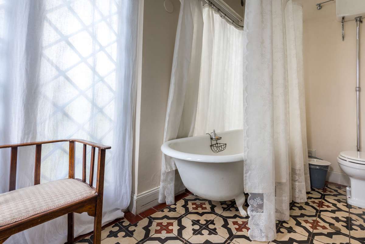 A clawfoot bathtub sits beneath a window with white curtains in a bathroom featuring patterned tile flooring and a chair.