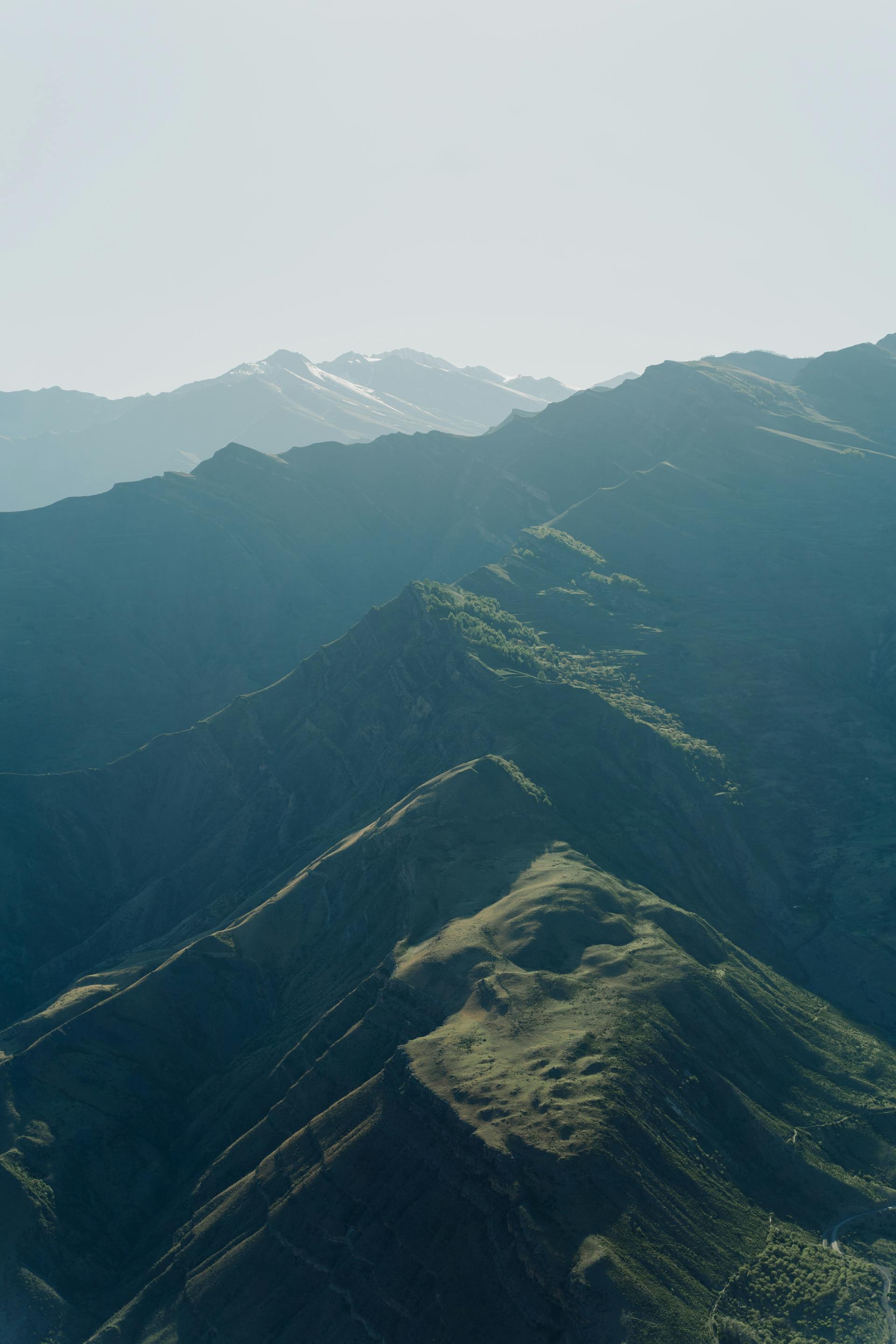 An aerial view of rugged, green mountain peaks and sharp ridges receding into a misty horizon under bright daylight.