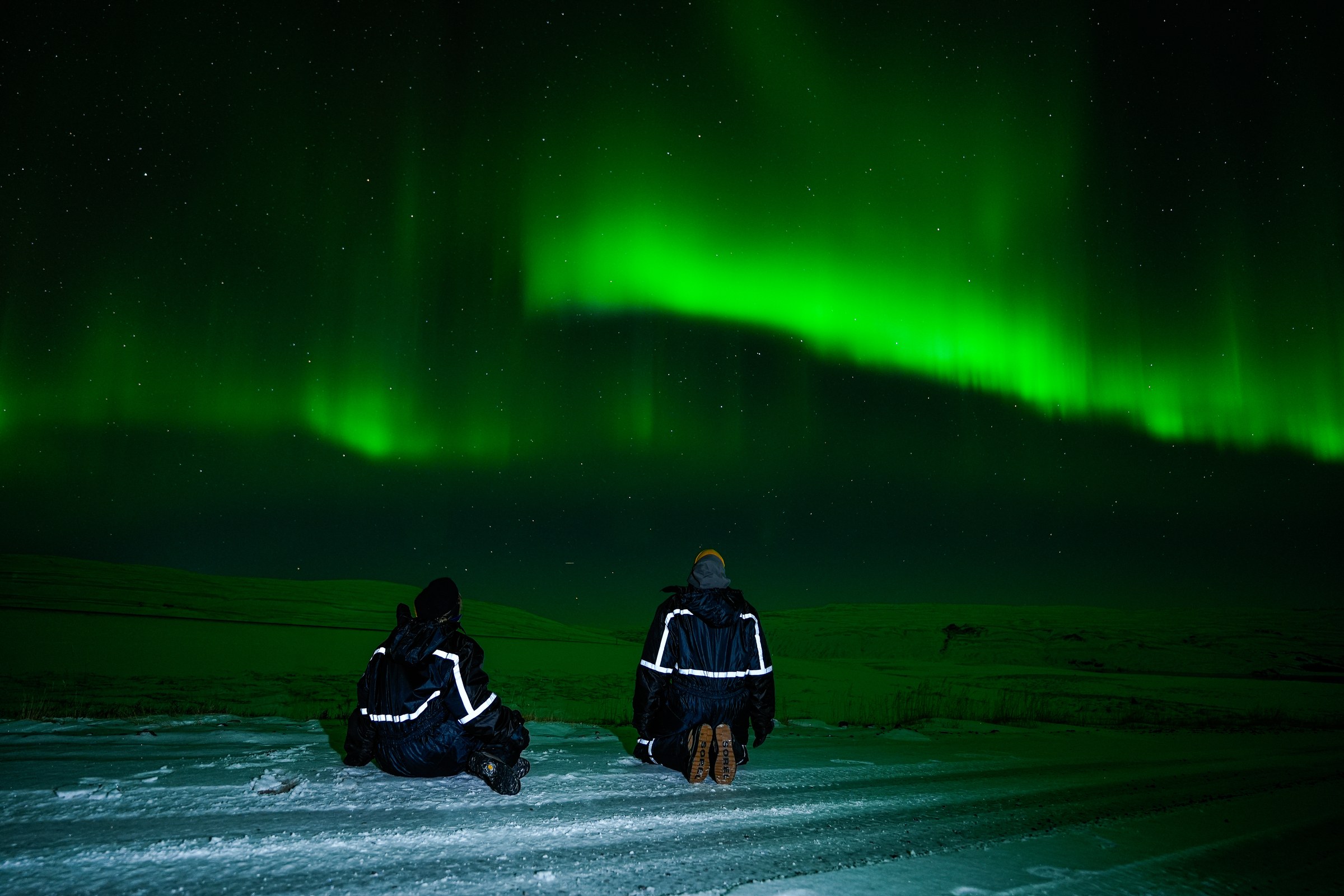 Two guests in snowsuits sitting on the ground watching a green aurora band low over the horizon
