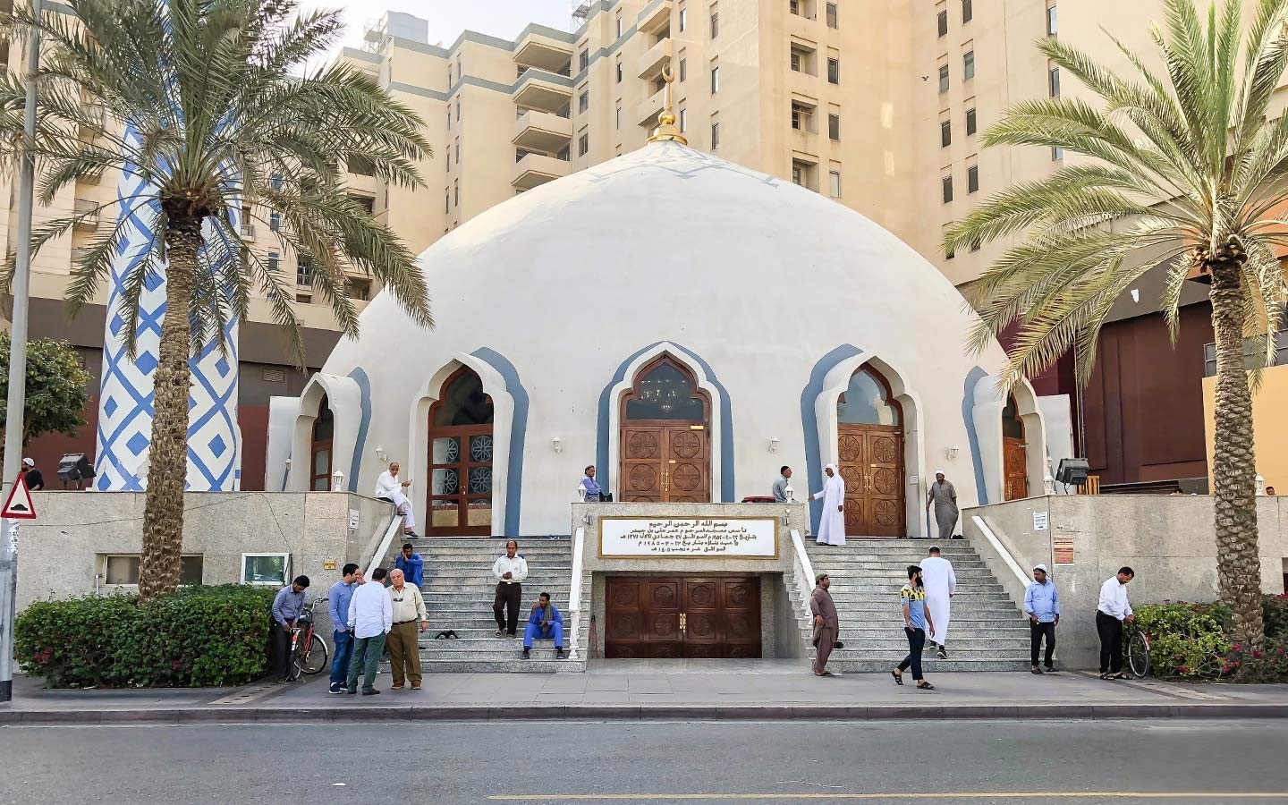 White domed mosque in Deira surrounded by palm trees and people gathered near its entrance on a sunny day.