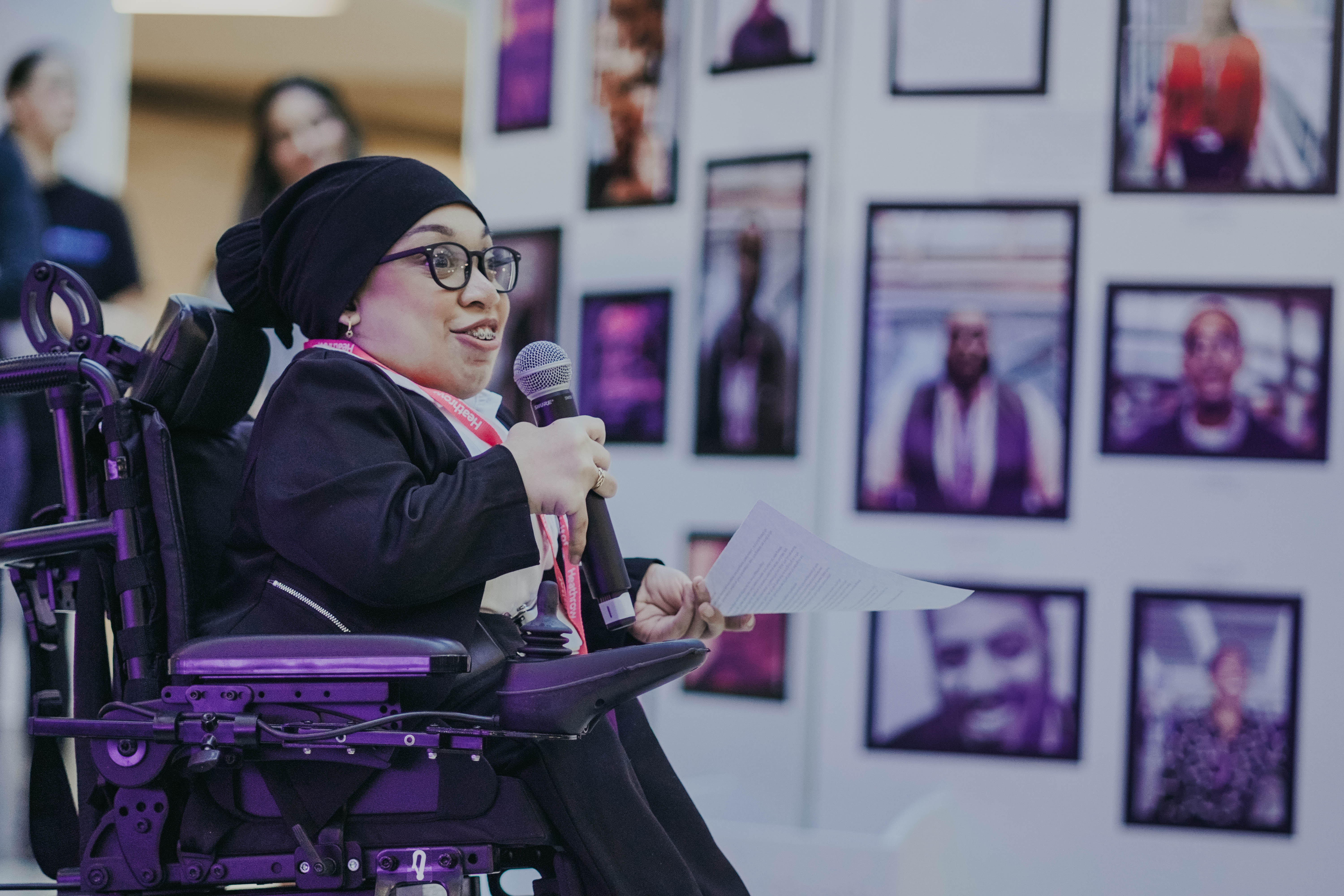 A woman in a motorized wheelchair, wearing a black hijab and glasses, speaks into a microphone while holding a paper. She is smiling and engaged, speaking in front of a wall decorated with numerous framed photographs.