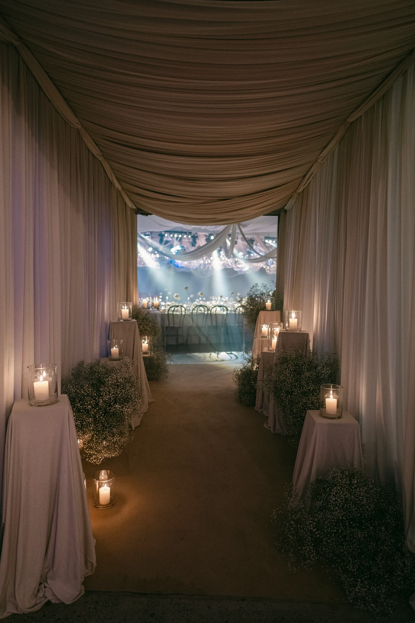 Pasillo inmersivo de boda con telas drapeadas en el techo, velas en fanales y nubes de gipsofila en el suelo.