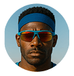 Close-up of a Black man with a blue headband, teal sunglasses, and a focused expression in a circular frame.