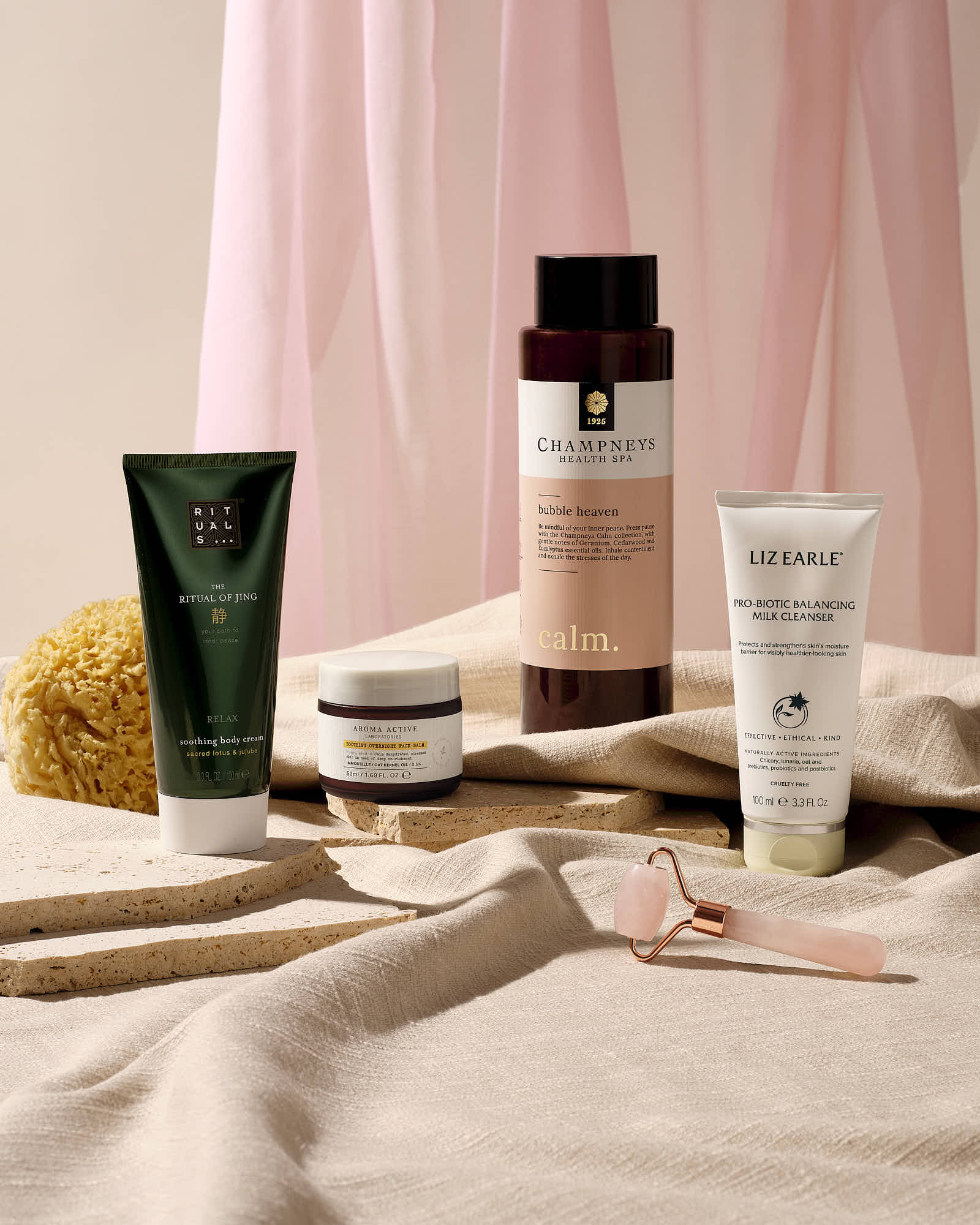 Spa essentials including cleansing milk, soothing body cream, bubble bath, and jade roller on fabric.