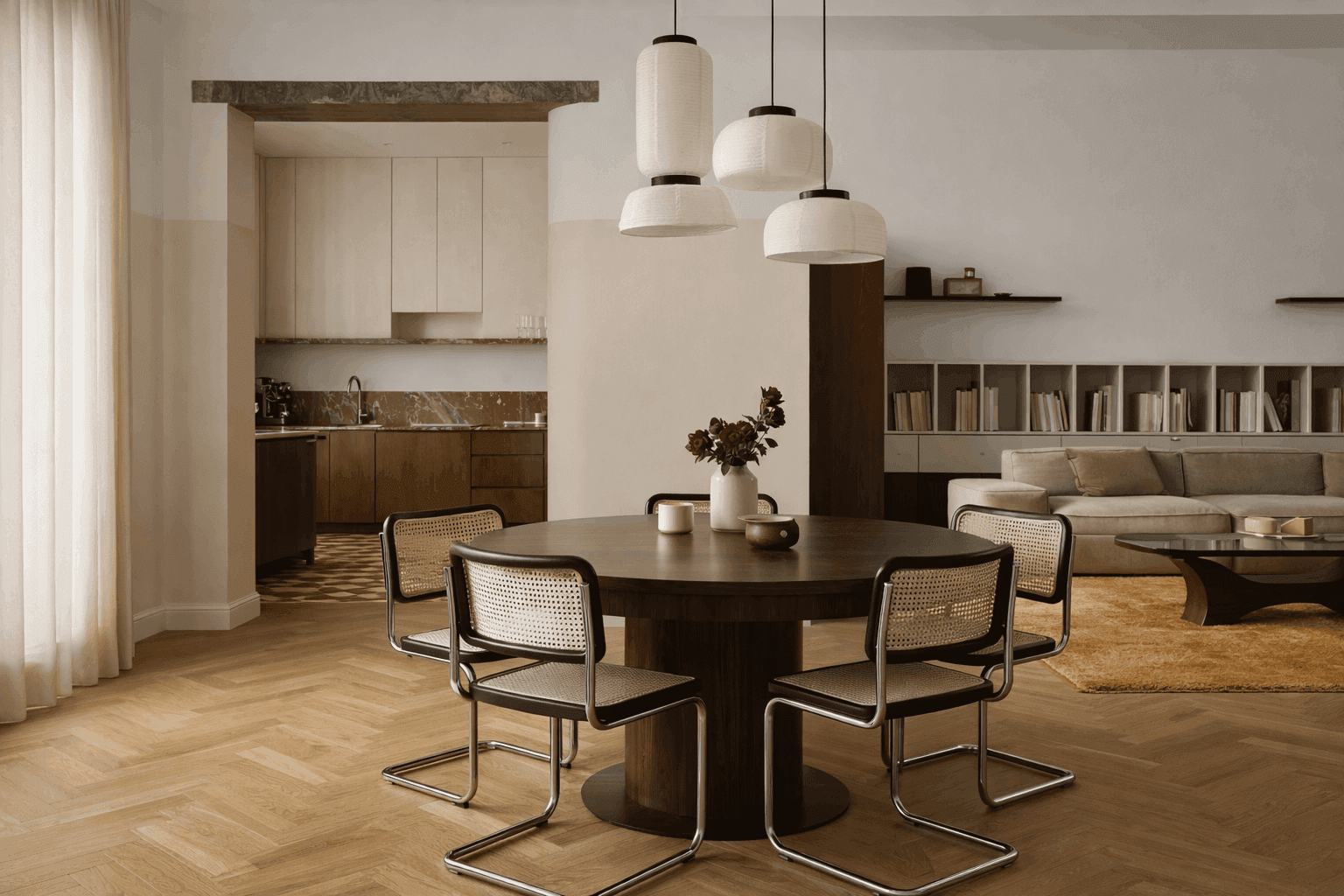 A modern, open-concept dining area features a round wooden table with four sleek, metal-framed chairs, illuminated by three pendant lights, against a backdrop of a cozy living room and a minimalist kitchen.