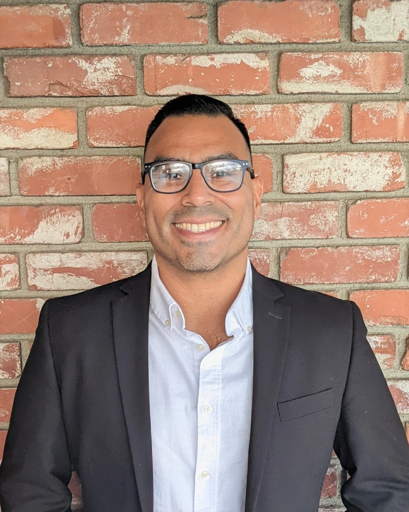 Uma pessoa usando óculos e um blazer preto formal sobre uma camisa branca está sorrindo diante de uma parede de tijolos vermelhos.
