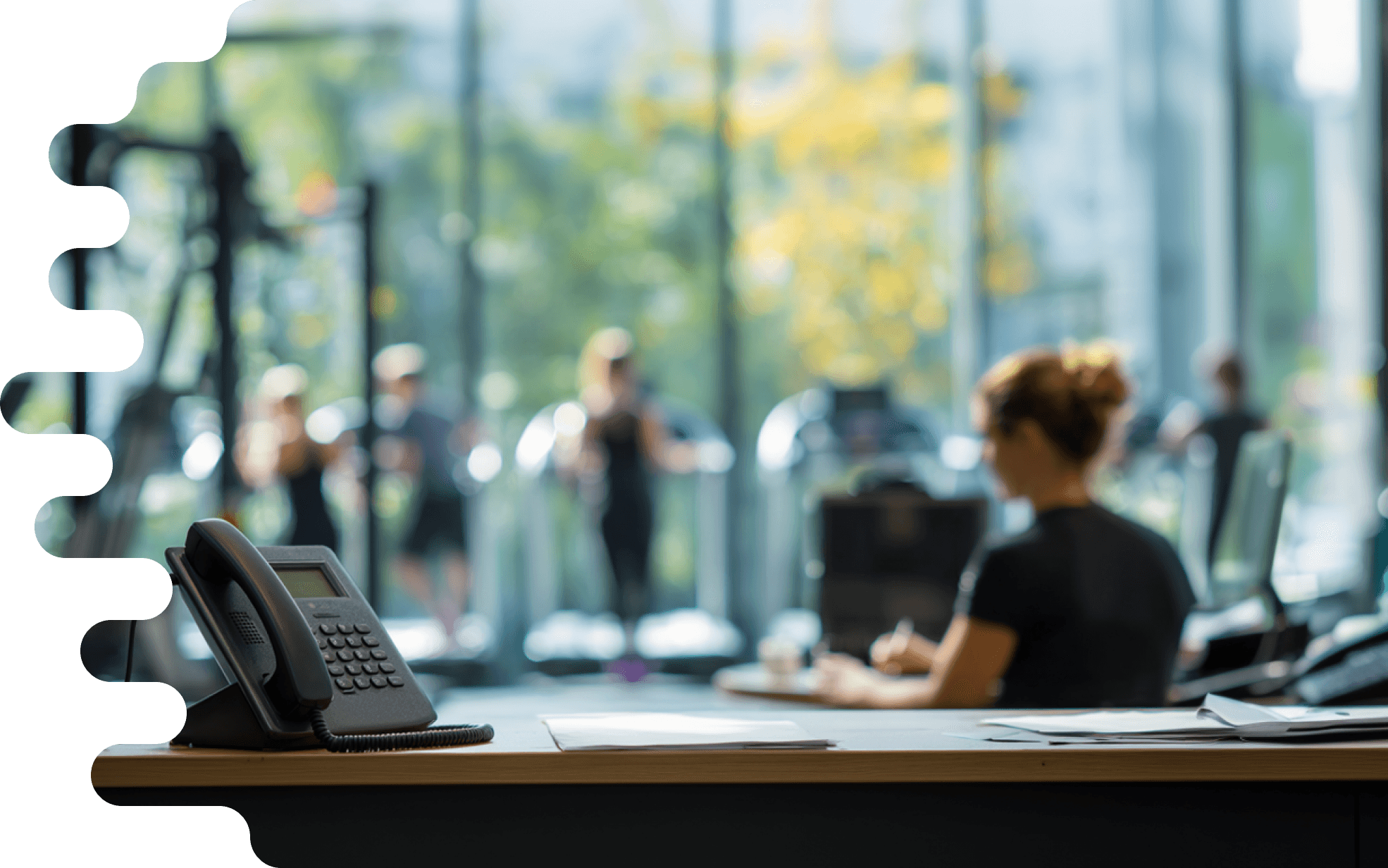 A sharply focused phone on the reception desk of a gym, next to a staff member at work. In the background, members are training out of focus. Symbolizes Safina AI phone assistant for professional accessibility in the studio.