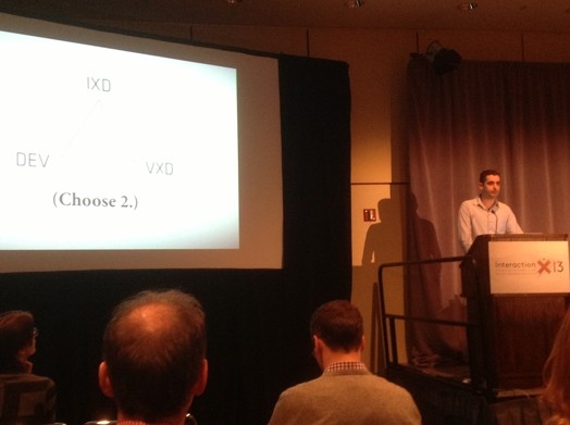 A photo of a man sitting at a stand and giving a lecture while showing something on a screen projector.
