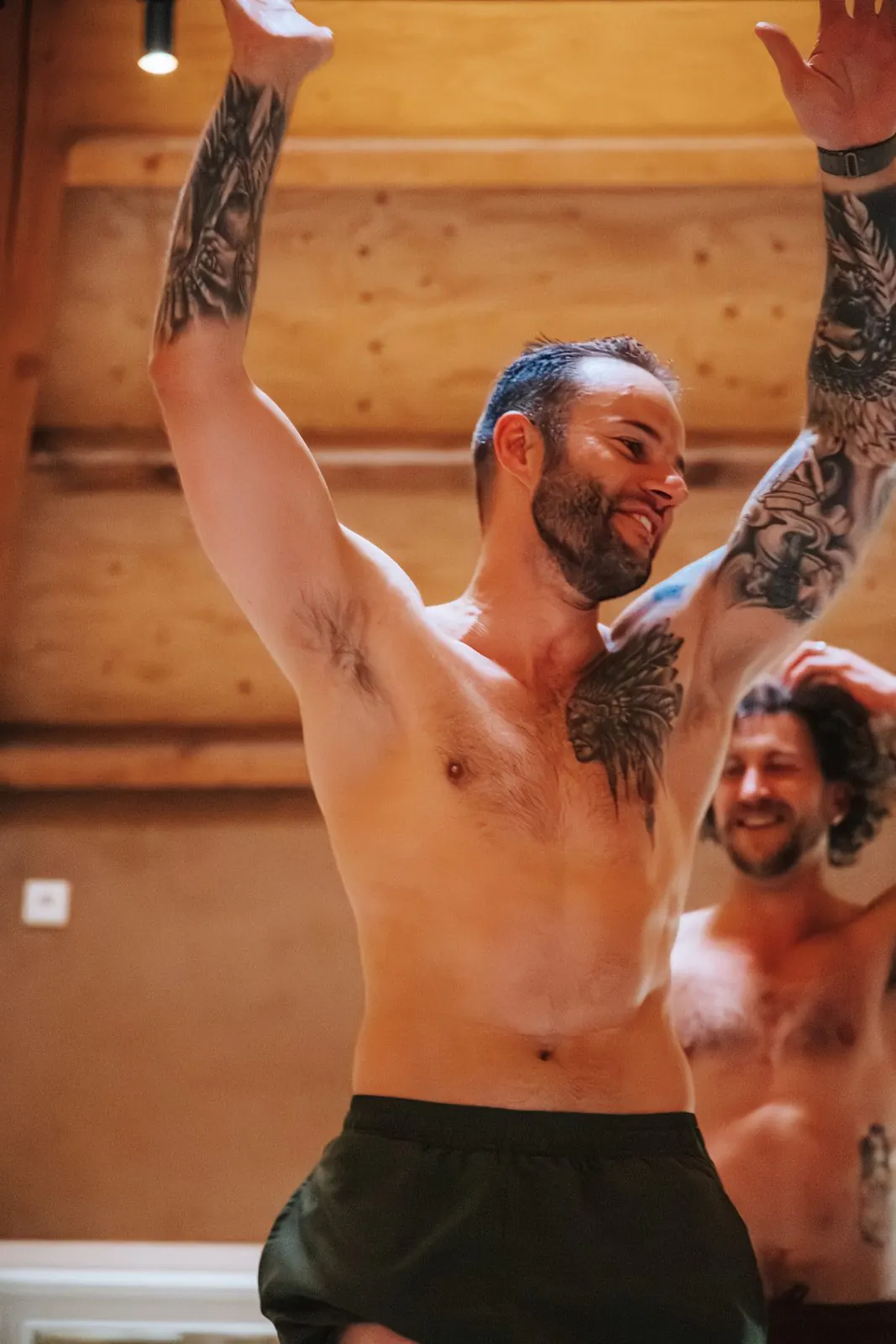 Een getatoeëerde man met ontbloot bovenlijf steekt lachend beide armen omhoog, achter hem lacht nog iemand