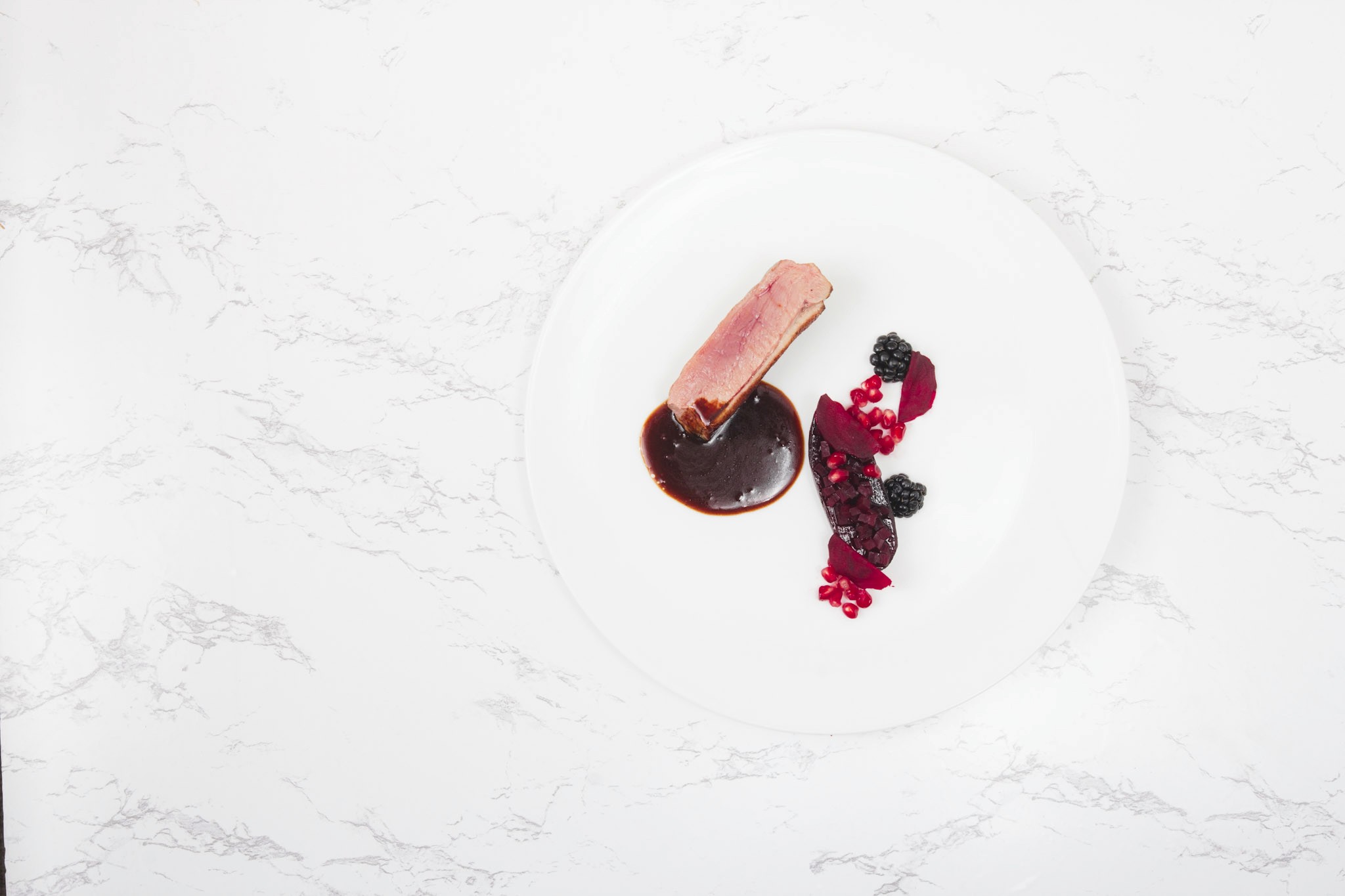 Top-down view of duck breast with blackberry sauce and a beetroot garnish, blackberries, and pomegranate seeds on a white plate.”