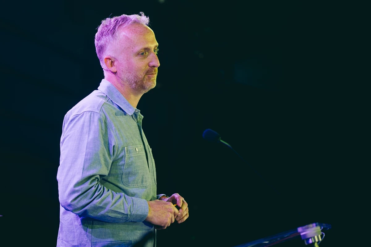 Tom Lawson standing on a stage under dramatic blue and purple lighting, wearing a light blue button‑up shirt. They are positioned near a lectern and microphone stand, holding their hands together as if mid‑talk. The dark background and spotlighting suggest a live presentation or speech at a professional event or conference.