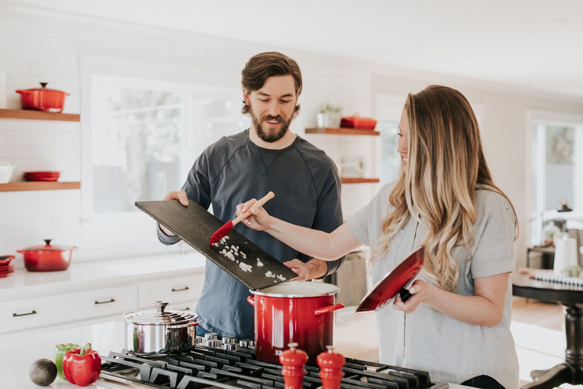 Een man en een vrouw zijn aan het kletsen in een lichte keuken, omringd door rood keukengerei en volop in interactie met elkaar.